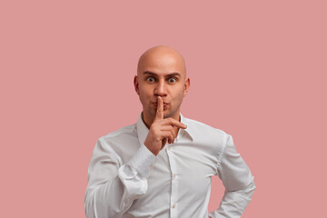 Be quiet please. Pleasant looking bald man with bristle, makes hush gesture, looks at the camera, asks for silence indoor. dressed in white shirt. Isolated over pink background.