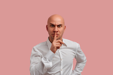 Shut up, please. Serious bald guy with bristle dressed in white shirt, says shh while shows hush gesture, dissatisfied with the noise from teenagers near. Isolated over pink background.