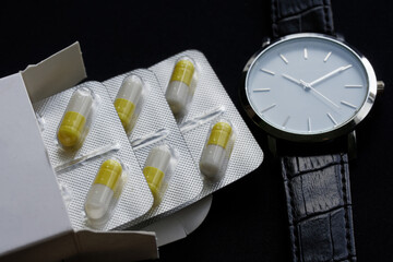 Yellow capsules in a silver blister and white cardboard box next to an elegant wristwatch. Schedule...
