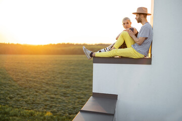 Stylish couple have a close conversation while sitting together on the rooftop with great sunset view on nature. Carefree lifestyle and romance concept. Wide view with copy space