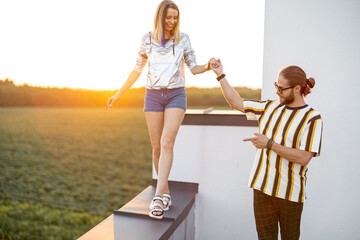 Stylish girl walks along the parapet of the roof with her boyfriend at sunset. Carefree lifestyle and romance concept