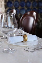 Festive table setting.A tablecloth with a textured pattern.Cotton napkin with a ring made of yellow metal.The concept of holidays.Wine glasses on table.White plates. Out of focus.Part of the interior.
