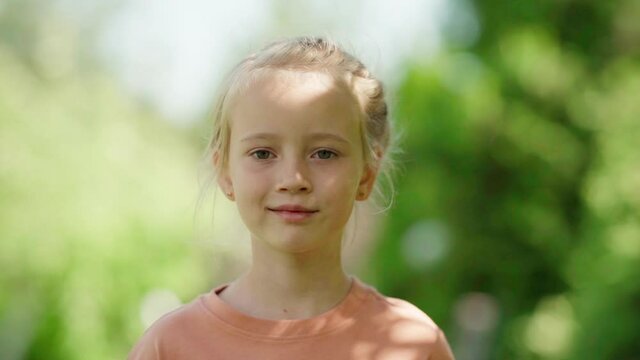 Slow motion portrait shot of cute blonde little girl looking at camera while posing outdoors on summer day, green foliage bokeh around