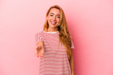 Caucasian blonde woman isolated on pink background stretching hand at camera in greeting gesture.