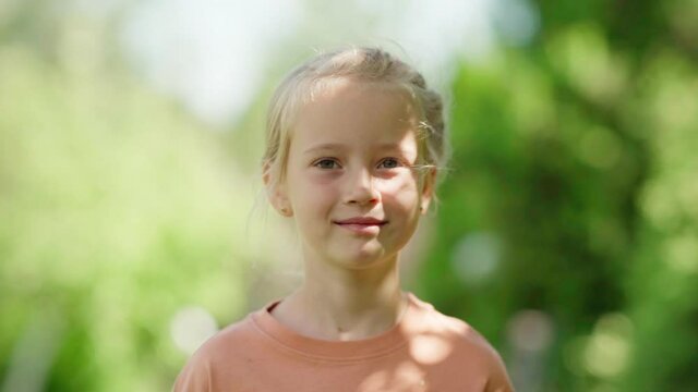 Slow motion portrait shot of beautiful blonde little girl looking away and forward while standing outside in park on summer day, green tree leaves bokeh around