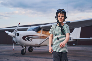 Adorable boy in aviation hat standing at aerodrome