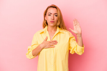 Caucasian blonde woman isolated on pink background taking an oath, putting hand on chest.