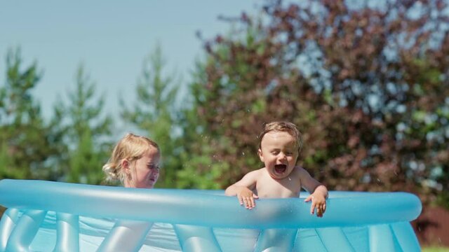 Little siblings having fun in inflatable swimming pool in backyard. Kids splashing water and screaming with excitement. Toddler boy leaning on poolside and jumping