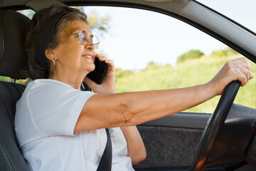 Happy mature woman wearing glasses driving car and talking on a cell phone. Smiling senior woman holding the wheel, road trip, active pensioner.