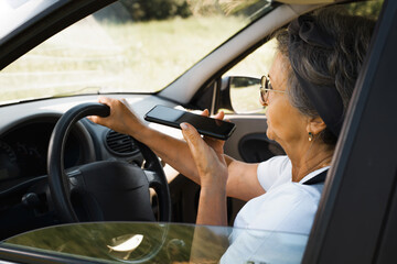 Senior woman with glasses talking on speakerphone on cell phone while driving. Elderly woman driving a car outside the city. Active retirement, road trip. Selective focus on face and smartphone