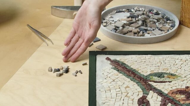 Male hands create mosaics floral pattern using tweezers and small pieces of granite stone. Master making handicraft in a creative workshop. Interesting hobby, art, work of art. Close up. Slow motion.