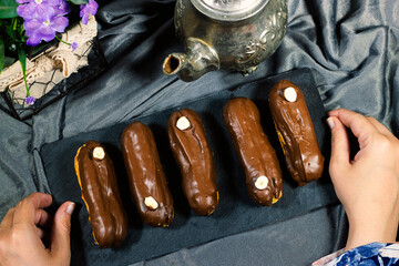 closeup hands woman holding french dessert eclairs with custard and chocolate icing decorated with hazelnuts and teapot, flowers pot