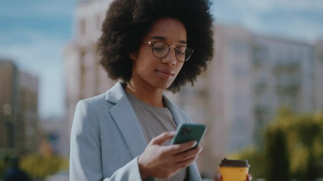 Afro-american modern young woman in business outfit and glasses chatting on smartphone screen and walking the city center street outside. Business girl.