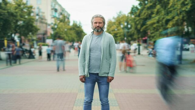 Zoom-in time lapse portrait of serious mature man standing alone outside in street with people rushing around. Modern society and life concept.