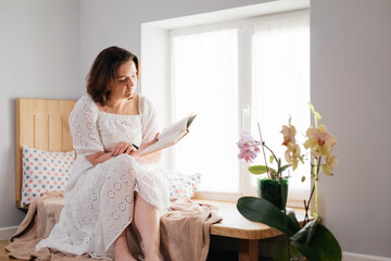 Mid age adult woman plus size reading a book by the window on the windowsill in the morning. Romantic cozy home atmosphere. Body positive and natural female beauty