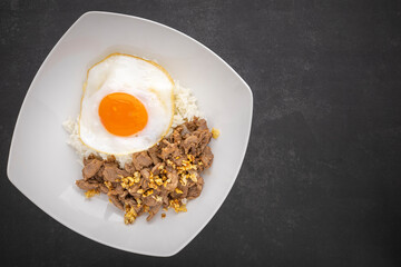Thai food, streamed rice topped with fried beef with garlic and fried egg  in ceramic plate on dark grey texture background with copy space for text