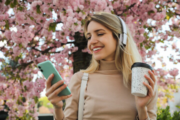 Happy woman with smartphone and coffee listening to audiobook outdoors on spring day