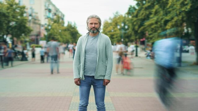 Zoom-out time lapse of gray-haired man in casual clothing standing outdoors in busy street and looking at camera with emotionless face. Society and life concept.