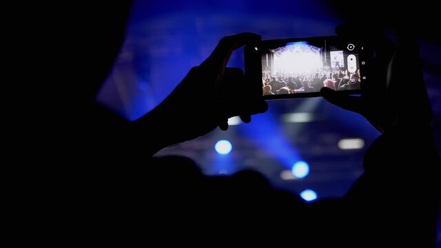Male Record Video on a Smart phone of a Night Rock Concert on Open Stage. Fan is shoot a video, photo on a mobile phone. Stage lighting with spotlights. Flickering light. Crowd of people on screen.