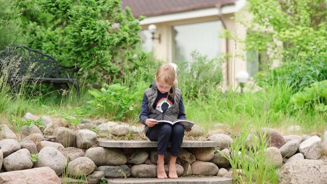 Cute little girl sitting on stone steps in house backyard and reading book aloud. Introvert kid spending summer vacation time in countryside