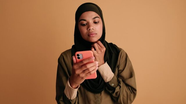 A positive arabian woman is correcting her burqa while looking at the camera of her smartphone standing isolated over beige background