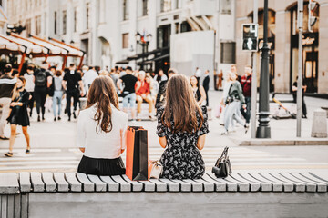 Two girls sitting on bench, summer day, view from behind, blurred pedestrians. Regular people out...