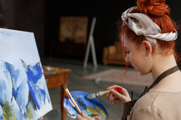 Talented female artist working on a painting in a dark creative studio. Beautiful creative girl sits in a workplace near a painted picture snowy mountains