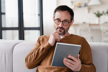 Pensive thoughtful middle-aged mature man using digital tablet for web browsing, online shopping, e-banking e-learning, working remotely as a freelancer.