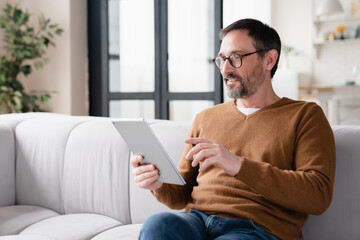 Relaxed mature middle-aged man using tablet, typing messages in social media, reading e-book,...
