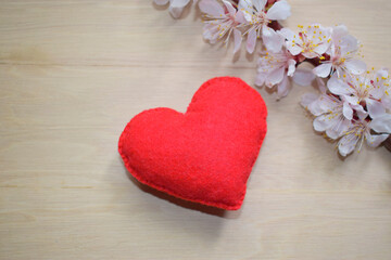 Red felt heart on a wooden surface, next to a blooming twig of a fruit tree.
