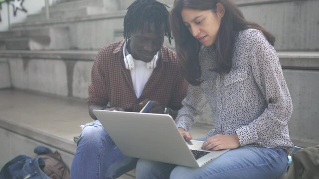 Two multiracial Freelance Colleagues or University Students working together in the City. African American man and Latin woman with laptop sitting on modern staircase
