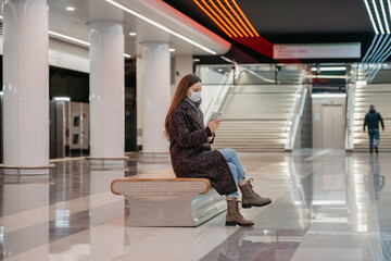 A woman in a medical face mask is sitting in the center of the subway platform and using a smartphone. A girl with long hair in a surgical mask is keeping social distance in the metro.