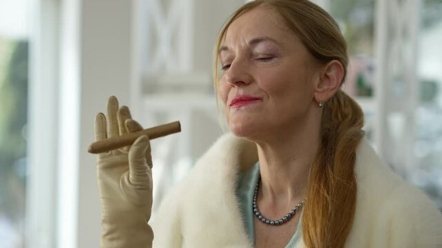Close-up portrait of stylish Caucasian middle aged woman smelling cigar smiling. Confident rich wealthy lady in gloves holding tobacco product standing indoors at luxurious home. Lifestyle and wealth