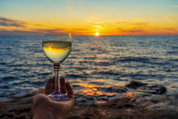 Human hand holding wine glass with sparkling wine against amazing sea landscape at sunset. Beautiful still life with alcohol drink on seascape background. Summer vacation, weekend at sea concept