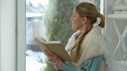 Elegant smart middle aged Caucasian woman looking out the window reading book at home indoors. Intelligent clever confident lady enjoying hobby on weekend at home. Lifestyle and happiness