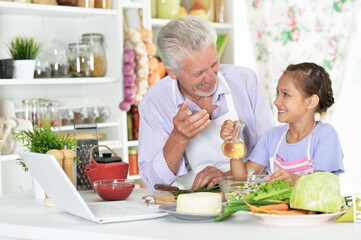 Portrait of senior man with granddaughter looking at laptop while preparing dinner in kitchen