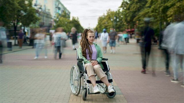Time lapse of disabled young woman with dreadlocks sitting in wheelchair in crowded pedestrian street and looking at camera. Youth and disability concept.