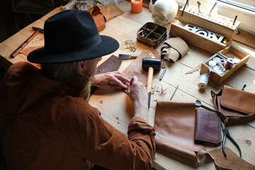 Obraz premium Rear view of shoemaker in hat sitting at the table using patterns and making shoes from leather in the workshop