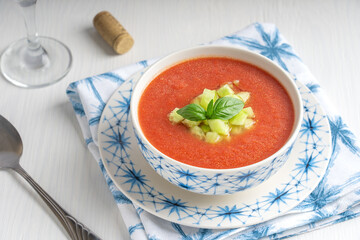 Gaspacho also called Andalusian gazpacho, cold refreshing soup made of raw, blended vegetables eaten during hot summers served in bowl with spoon and glass of wine on white wooden background on towel