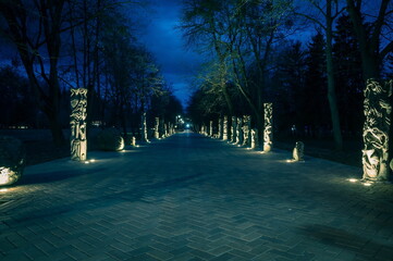 The tiled road in the night park with stone idols illuminated by lamps. Stone statues in the park...