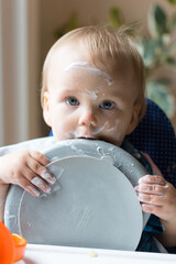13 month old baby holding plate and licking it after eating yogurt; messy face and hands as she self feeds