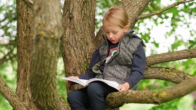 Tilt up of cute barefoot little girl sitting on tree branch in park or backyard and reading book aloud. Introvert kid spending summer time alone