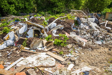 Construction debris is scattered in the backyard of the building. Various rubbish lies on the grass in a big city. Old boxes, plywood and plastic bags on a background of green trees