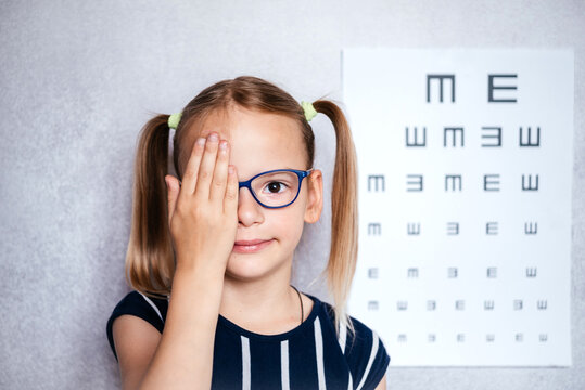 Little girl wearing eyeglasses taking eyesight test before school with blurry eye chart at the background