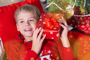 Obraz na płótnie Canvas Smiling Blond Boy Holding Festive Gift Box and Lying on the floor near Christmas Tree