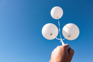 A male hand holds three white air balloons against a very blue sky.
