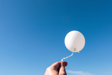 The fingertips of a male hand holds a white air balloon against a very blue sky.


