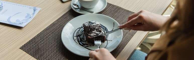 Cropped view of woman cutting dessert near cup and menu in cafe, banner.