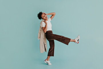 Excited african american woman in pants dancing on turquoise background. Full length view of adorable black girl holding trench coat.