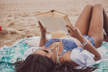 Vertical camera position of a black girl lying on the beach holding a book while looking forward on a sunny day. Summer vibes concept 2021.
